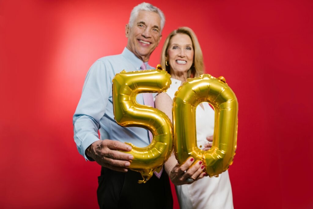 Happy senior couple celebrating 50th anniversary with golden balloons against a red studio backdrop.